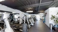 This fitness center offers a comprehensive workout environment spread across a single level. The space features an industrial-style exposed ceiling with visible ductwork, pipes, and overhead fluorescent lighting, lending the gym a utilitarian, no-frills aesthetic. Large mirrored walls line one side of the room, visually expanding the space and allowing users to monitor their form during workouts.

The cardio area is well-stocked with a row of treadmills and elliptical machines, all of modern design and arranged to maximize floor space efficiency. Additional equipment, including stationary bikes and step machines, rounds out the cardio offerings. Further into the space, a designated stretching and free weights area is visible, complemented by a stability ball and exercise mats. A wall-mounted flat-screen television is positioned for easy viewing from the cardio equipment.

Natural light filters in through windows along one wall, brightening the space alongside the overhead lighting. The flooring is durable and suitable for high-traffic fitness use. Overall, the gym provides a functional and well-organized environment catering to a variety of fitness needs.