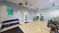 Located at Madison Place in Kernersville, NC, this single-floor fitness center is thoughtfully designed to support a full range of workout routines. The space features light wood-tone flooring throughout, complemented by soft blue-gray walls and bright linear lighting paired with ceiling fans that keep the environment comfortable during exercise sessions.

The cardio zone is equipped with multiple treadmills, elliptical trainers, and a stair climber, all positioned near large windows with slatted blinds that allow natural light to filter in. Mounted flat-screen televisions above the cardio equipment provide entertainment during workouts. Adjacent to this area is a multi-station cable strength machine and a dedicated free-weight section featuring a full rack of dumbbells in varying sizes, along with a weight bench and rubber floor mats for safe lifting.

Toward the opposite end of the room, stationary cycling bikes provide an additional cardio option in a more open area of the floor. A wall of mirrors expands the sense of space and assists with form checks during exercise. Wall-mounted accessories, including resistance bands and medicine balls, add further versatility to the training options available.

The space is well-ventilated with multiple ceiling fans and includes a glass-paned entry door. Overall, the fitness center offers a clean, well-organized environment suited for a variety of fitness goals.