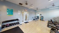 Located at Madison Place in Kernersville, NC, this single-floor fitness center is thoughtfully designed to support a full range of workout routines. The space features light wood-tone flooring throughout, complemented by soft blue-gray walls and bright linear lighting paired with ceiling fans that keep the environment comfortable during exercise sessions.

The cardio zone is equipped with multiple treadmills, elliptical trainers, and a stair climber, all positioned near large windows with slatted blinds that allow natural light to filter in. Mounted flat-screen televisions above the cardio equipment provide entertainment during workouts. Adjacent to this area is a multi-station cable strength machine and a dedicated free-weight section featuring a full rack of dumbbells in varying sizes, along with a weight bench and rubber floor mats for safe lifting.

Toward the opposite end of the room, stationary cycling bikes provide an additional cardio option in a more open area of the floor. A wall of mirrors expands the sense of space and assists with form checks during exercise. Wall-mounted accessories, including resistance bands and medicine balls, add further versatility to the training options available.

The space is well-ventilated with multiple ceiling fans and includes a glass-paned entry door. Overall, the fitness center offers a clean, well-organized environment suited for a variety of fitness goals.