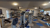 This single-floor fitness center is thoughtfully designed to accommodate a wide range of workout needs. The main exercise area features a robust collection of Hoist-brand strength machines, including cable systems, leg press, and various resistance stations, providing users with a thorough strength training circuit. A dedicated free weights section lines one wall, equipped with kettlebells, resistance bands, a weight rack, and dumbbells organized for easy access.

The cardio zone is outfitted with True-brand treadmills and additional cardio equipment, positioned near windows that allow natural light to fill the space. Multiple wall-mounted flat-screen televisions are strategically placed throughout the facility, keeping members entertained during workouts.

Large wall mirrors expand the sense of space and allow exercisers to monitor their form. Ceiling fans are installed throughout to promote air circulation and comfort. A separate alcove with wood-look flooring serves as a functional stretching and flexibility area, stocked with stability balls and foam rollers, and features a built-in water fountain for easy hydration.

The flooring throughout the main workout area consists of a visually distinctive hexagonal tile pattern in blue, gray, and tan tones. Recessed and flush-mount lighting, combined with natural light from multiple windows, ensures the space is well-lit and inviting.