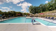 Located at 210 Highland Corporate Dr in Cumberland, Rhode Island, this outdoor recreational facility offers a well-appointed pool area designed for both leisure and accessibility. The centerpiece is a large, sparkling swimming pool with clear blue water, complemented by a separate, smaller wading or spa section. A dedicated accessibility lift is installed poolside, ensuring inclusive use of the facility for all visitors.

The generous concrete pool deck surrounds the water on all sides, providing ample space for lounging. Multiple patio umbrellas in deep red and neutral tones are positioned throughout, offering shaded seating areas with outdoor tables and chairs for relaxation and socializing. The pool area is fully enclosed by a black iron security fence, ensuring safety while maintaining open sightlines to the surrounding greenery.

Adjacent to the pool, a well-maintained outdoor grilling and recreation zone features a gas grill station set within a beautifully landscaped area. Flowering hydrangeas, ornamental shrubs, and manicured lawn borders bring vibrant color and natural charm to the space. A rocky hillside and mature tree line form a scenic natural backdrop. The building exterior features classic white vinyl siding with a red brick base, and a dedicated pool equipment room is accessible directly from the deck area.