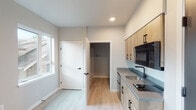 This single-floor unit showcases a clean, modern design with a neutral color palette and light-toned luxury vinyl plank flooring that runs throughout the space, lending a cohesive and contemporary feel. The kitchenette is efficiently designed, featuring natural wood-toned cabinetry with matte black hardware, a gray quartz-style countertop, an undermount sink with a matte black faucet, a built-in cooktop, and an over-the-range microwave. A full-size refrigerator is positioned adjacent to the cabinetry, maximizing functionality within the compact kitchen footprint. The open living area benefits from natural light provided by several windows, offering views of the surrounding neighborhood. Walls are finished in soft gray tones, complementing the bright white trim and doors with black hardware found throughout. A dedicated utility area houses the electrical panel and additional access points, ensuring practical infrastructure is neatly integrated. The unit also includes a walk-in storage or utility closet accessible from the main living space. Recessed lighting provides clean, even illumination, while HVAC venting is discreetly incorporated into the walls. Overall, the space presents a fresh, move-in-ready environment with functional, well-considered detailing.