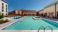 This contemporary apartment complex in Suwanee, GA, offers a blend of sophistication and comfort. The architecture combines light-colored brick facades with modern siding, creating an appealing visual contrast. Large windows and private balconies characterize the building's exterior, maximizing natural light and outdoor views for residents.

The property boasts an impressive array of amenities. A centerpiece is the resort-style pool, featuring a zero-entry design, in-water lounge chairs, and surrounded by ample deck space with modern poolside furniture. Adjacent to the pool area, residents can enjoy outdoor lounge spaces with comfortable seating, fire pits, and string lighting for ambiance.

Landscaping plays a crucial role in the complex's design, with manicured lawns, mulched plant beds, and a variety of trees and shrubs enhancing the outdoor spaces. Several courtyard areas offer additional recreational options, including a putting green, hammocks, and outdoor dining areas with grills.

For fitness enthusiasts, the complex provides outdoor exercise equipment and open green spaces suitable for yoga or other activities. The property also includes covered parking options and appears to be situated near wooded areas, offering a balance of urban convenience and natural surroundings.

Inside, the apartments likely feature modern finishes and open-concept layouts, as suggested by the contemporary exterior design. Overall, this apartment complex presents a high-end living experience with a focus on outdoor amenities and community spaces, catering to those seeking a luxurious, active lifestyle in a suburban setting.