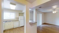 This single-story space offers a practical and open layout that connects the kitchen and living areas seamlessly. The kitchen is equipped with white cabinetry, a full-size refrigerator, a dishwasher, and a countertop workspace, all set against light-toned surfaces that contribute to a clean, bright aesthetic. A skylight overhead enhances the natural light in the kitchen, making the space feel airy and welcoming. A half-wall peninsula separates the kitchen from the adjoining living area, providing a functional transition between the two spaces while maintaining an open sightline throughout. The living area features warm wood-style flooring that runs continuously, adding cohesion and a sense of warmth to the interior. Vertical blinds cover a sliding glass door in the living area, allowing for adjustable natural light and potential access to an outdoor space. Neutral yellow-toned walls and white trim throughout give the space a consistent and understated palette. Ceiling vents indicate a central HVAC system for year-round climate comfort. Overall, the layout is efficient and well-lit, suited for everyday living.