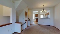 This two-story home offers a well-organized and functional layout designed for comfortable everyday living. The main level opens into a spacious carpeted living area that flows seamlessly into the adjoining spaces, creating an inviting and connected atmosphere. A classic wood-railed staircase with warm tones serves as a central architectural feature, providing access to the upper level while adding a traditional charm to the interior.

The living area transitions naturally into the kitchen and dining space, framed by a wide open archway. The kitchen is outfitted with warm wood cabinetry and a dark countertop, complemented by a white refrigerator and a window that brings in natural light. A raised bar-height counter separates the kitchen from the adjacent area, ideal for casual dining or entertaining. Overhead, an elegant multi-arm chandelier with warm-toned bulbs adds a refined focal point to the dining space.

Throughout the main level, the neutral white walls and light-toned flooring create a bright and airy ambiance. The upper level, accessible via the staircase, features a vaulted ceiling that enhances the sense of spaciousness and openness. Overall, this home combines traditional design elements with a practical, flowing layout suited for a variety of lifestyles.