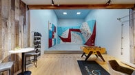 Located at 1720 Kirkwood Dr in Fort Collins, Colorado, this distinctive single-floor space blends active recreation with a thoughtfully designed lounge environment. The centerpiece of the space is a large, multi-panel indoor bouldering wall, featuring dynamic geometric sections in red, blue, and white with colorful climbing holds throughout. The wall spans two connected surfaces, offering a variety of routes and angles, and is complemented by padded crash mats below for safety. Track lighting and recessed ceiling fixtures ensure the climbing area is well-illuminated throughout.

Adjacent to the climbing wall, the activity zone includes a full-size foosball table, adding to the recreational atmosphere. A dedicated shoe rack provides organized storage for climbing footwear. The lounge area transitions seamlessly from the active zones, anchored by a striking full-height reclaimed wood accent wall that introduces warmth and rustic character. Light hardwood flooring runs throughout, unifying the space. The lounge is furnished with a bistro-height table, modern bar stools, and a comfortable accent chair. Exposed wooden ceiling beams and industrial-style track lighting contribute to the space's contemporary, loft-inspired aesthetic. A glimpse of an upper mezzanine level adds vertical depth and architectural interest to the overall design.