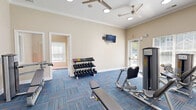 Located at 221 Fairforest Way in Greenville, SC, this well-appointed fitness center offers a comprehensive workout environment across a single, open floor. The space is finished with vibrant blue multi-tone carpet tiles that lend a modern, energetic feel to the room. Warm beige walls adorned with crown molding provide a clean, polished backdrop, while recessed lighting and multiple ceiling fans ensure the space remains bright and comfortable during use. Large windows fitted with horizontal blinds line the walls, inviting natural light and offering views of the outdoor pool area, creating an airy and motivating atmosphere. The fitness center is equipped with a wide range of exercise machines, including treadmills, ellipticals, stationary bikes, and a variety of strength-training and cable machines. A dedicated free weights section features a multi-tiered dumbbell rack alongside weight benches, catering to a full range of strength training needs. A wall-mounted television provides entertainment during workouts. Additional amenities include a compact storage counter with a mini-refrigerator, posted fitness center rules, and a mirrored wall section that enhances the sense of space. The layout flows openly between cardio and strength zones, making the facility both functional and easy to navigate.