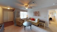 This single-floor residence offers a thoughtfully laid-out interior with a warm and comfortable ambiance throughout. The living room serves as the central gathering space, featuring plush carpeting, a ceiling fan with light fixture, and a large window that invites natural light and outdoor views. The room comfortably accommodates a full seating arrangement, making it ideal for both relaxing and entertaining. The kitchen is equipped with white cabinetry, an electric range, an over-the-range microwave, and dark accent walls that lend a bold contrast to the bright appliances. A dining area is visible just beyond, offering a convenient flow for daily living. The residence includes multiple bedrooms, each carpeted and fitted with ceiling fans. The bedrooms feature windows with natural light, and at least one includes a walk-in closet. The rooms offer ample space for full bedroom furniture sets. Both bathrooms follow a consistent design with wood-look flooring, white vanity cabinetry, a single-sink countertop, a large mirror, and a shower-tub combination enclosed by a curtain. The neutral color palette and functional layouts throughout the home create a cohesive, livable environment suited for a variety of lifestyles.
