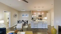 This single-floor residence showcases a thoughtfully designed open-concept layout that integrates the living, dining, and kitchen areas into one cohesive, flowing space. The kitchen is a standout feature, outfitted with light flat-panel cabinetry, stainless steel appliances including a refrigerator and microwave, and a sleek peninsula with a white quartz-style countertop. Three stylish pendant lights hang above the peninsula, providing focused task lighting while adding a modern decorative touch. Bar-height seating along the peninsula creates a casual dining option in addition to the dedicated dining area nearby. The dining space comfortably accommodates a full table and chairs, framed by framed wall art that adds a curated, welcoming feel. The living area flows naturally from the dining space, offering an inviting retreat anchored by soft furnishings. Rich wood-tone flooring runs throughout, providing warmth and continuity across all areas. The ceilings are generously proportioned, with recessed and track lighting ensuring the space remains bright and well-lit. Neutral wall tones create a versatile backdrop, complementing both the light cabinetry and the darker flooring. Overall, the space presents a clean, contemporary aesthetic with a practical and comfortable layout ideal for everyday living and entertaining.
