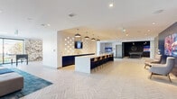 Located at 770 Brookfield Rd in Ottawa, Ontario, this generously sized amenity lounge spans a single open-concept floor, offering a well-appointed communal space designed for both relaxation and entertainment. The layout flows seamlessly across distinct functional zones, all unified by a striking light-toned herringbone tile floor and crisp white ceilings with recessed lighting throughout.

At the heart of the space sits a long white island bar with dark navy cabinetry, pendant lighting, and bar seating — ideal for social gatherings. A decorative tile backsplash and wall-mounted television add to the polished aesthetic. Adjacent to the bar, a lounge area features a large sectional sofa set atop vibrant teal area rugs, accompanied by an elegant linear fireplace framed in marble-look tile and a large wall-mounted display screen flanked by lush vertical living wall installations, bringing a refreshing natural element to the interior.

The game zone is distinctly defined by dramatic dark steel-framed partitions with a black ceiling, pendant Edison bulbs, and a "Game On" neon sign. This area houses a ping pong table and a pool table, creating an energetic entertainment hub. Floor-to-ceiling windows invite natural light and outdoor views into the space. A foosball table and additional casual seating round out the amenities, while a large-scale artistic mural adds a creative, curated character to the overall environment.