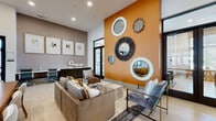 This thoughtfully designed single-floor amenity space blends bold aesthetics with versatile functionality across several distinct zones. The entryway lounge immediately commands attention with its striking burnt-orange accent wall adorned with an eclectic gallery of decorative mirrors in varied shapes and textures, setting a vibrant, contemporary tone. Light wood-look tile flooring flows throughout, tying the open layout together seamlessly.

A formal gathering area features a substantial wood dining table surrounded by upholstered chairs beneath an angular statement chandelier, making it ideal for meetings or social events. Adjacent to this, a comfortable lounge area provides plush seating and a wall-mounted television against a bold horizontally-striped orange, white, and gray accent wall, with large floor-to-ceiling windows and glass doors that bathe the space in natural light and offer outdoor views.

The bar and kitchen area stands out with a long white countertop island lined with tall upholstered barstools, dramatic pendant lighting in white and gold, and a textured mosaic accent wall hosting an additional television. A built-in coffee station adds a practical amenity touch. Tucked nearby, a private nook with a cozy booth-style banquette and framed artwork provides an intimate retreat. The space also features a billiards table and workstation area, completing a well-rounded, multi-use environment filled with curated décor and lush indoor greenery.
