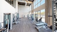Located at 10900 Legacy Gateway Cir in Fort Myers, FL, this expansive fitness center is designed to deliver a comprehensive workout experience across a single, well-appointed floor. The facility is organized into distinct zones, each tailored to a specific type of training. The strength training area commands attention with soaring double-height ceilings, warm wood-accented beam details, and dramatic floor-to-ceiling windows that flood the space with natural light and offer views of the surrounding landscape. A wide array of strength machines and benches are thoughtfully arranged throughout this open area. The cardio zone features a row of treadmills, stationary bikes, and other aerobic equipment positioned directly along expansive window walls, providing natural light and outdoor views of the palm-lined surroundings. An additional area with an exposed concrete ceiling and industrial-style track lighting houses stair climbers, a dumbbell rack, mirrored walls, and functional training equipment. A dedicated studio room with warm hardwood flooring, high ceilings, a large wall-mounted display, and full-length mirrors provides an ideal space for cycling, yoga, and group fitness classes. Kettlebells, medicine balls, and stability equipment complete the studio's offerings. Hallways connect the various zones, creating a cohesive and functional flow throughout the entire facility.