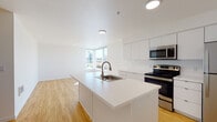 Located at 20433 SE Stark St in Gresham, Oregon, this modern single-floor unit offers a clean, contemporary design throughout. The open-concept layout seamlessly connects the kitchen and living area, creating a bright and functional living space. Large windows flood the interior with natural light and frame views of the surrounding Pacific Northwest landscape, including mature evergreen trees.

The kitchen is well-appointed with crisp white cabinetry extending to the ceiling for ample storage, a central island with a built-in sink and dishwasher, and a full suite of stainless steel appliances including a refrigerator, range, microwave, and dishwasher. Light bamboo-style wood flooring runs throughout the main living areas, adding warmth to the minimalist white interior.

The bedroom features neutral carpet flooring, a ceiling fan, and a window that brings in natural light. A walk-in closet provides generous storage. The unit includes two bathrooms: a full bath with a soaking tub and shower combination, and a half bath with a white vanity and sink. Both bathrooms continue the clean, modern aesthetic with light flooring and white finishes. The overall ambiance is bright, airy, and move-in ready.