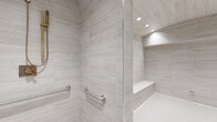 This single-floor space showcases a meticulously designed bathroom that combines elegance with accessibility. The walk-in shower is a standout feature, clad floor-to-ceiling in large-format, light gray stone-look tiles that lend a clean, contemporary aesthetic throughout. A frameless glass panel defines the shower enclosure, maintaining an open and airy feel while recessed ceiling lighting bathes the space in a warm, even glow. Built into the shower is a tiled bench seat, offering a comfortable and seamlessly integrated resting area, complemented by a matching tiled niche shelf ideal for storage. Polished stainless steel grab bars are thoughtfully positioned along the walls, providing both safety and a sleek, modern accent. The floor transitions to a fine mosaic tile, adding subtle texture underfoot while enhancing slip resistance. The overall design palette of soft whites and cool grays creates a serene, spa-like ambiance, while the careful attention to accessible design elements ensures the space is both functional and welcoming for all users.