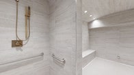 This single-floor space showcases a meticulously designed bathroom that combines elegance with accessibility. The walk-in shower is a standout feature, clad floor-to-ceiling in large-format, light gray stone-look tiles that lend a clean, contemporary aesthetic throughout. A frameless glass panel defines the shower enclosure, maintaining an open and airy feel while recessed ceiling lighting bathes the space in a warm, even glow. Built into the shower is a tiled bench seat, offering a comfortable and seamlessly integrated resting area, complemented by a matching tiled niche shelf ideal for storage. Polished stainless steel grab bars are thoughtfully positioned along the walls, providing both safety and a sleek, modern accent. The floor transitions to a fine mosaic tile, adding subtle texture underfoot while enhancing slip resistance. The overall design palette of soft whites and cool grays creates a serene, spa-like ambiance, while the careful attention to accessible design elements ensures the space is both functional and welcoming for all users.