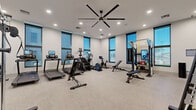 This fitness center is thoughtfully designed to provide a complete workout environment within a generously sized, open-plan layout. The space features light-toned speckled flooring throughout, offering durability and easy maintenance, while smooth white walls are accented by a bold navy blue feature wall that adds a modern, energetic character to the room.

Recessed lighting is distributed evenly across the ceiling, supplemented by a large ceiling fan that ensures consistent air circulation throughout the space. Multiple tall, dark-framed windows line the walls, allowing natural light to filter in while providing exterior views, contributing to an open and airy atmosphere.

The cardio zone is well-stocked with several treadmills equipped with individual digital displays, a stair climber, an elliptical trainer, a stationary bike, and a rowing machine. The strength training area includes a multi-functional cable machine, a squat rack, adjustable benches, and a neatly organized dumbbell rack. A functional training station offers resistance bands and kettlebells for diverse workout options.

A full-length mirror wall on one side enhances the sense of space and supports proper form during exercise. The overall design balances functionality with a clean, contemporary aesthetic suited to a professional fitness environment.