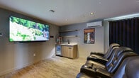 Located at 1946 Ambrosi Rd in Kelowna, BC, this dedicated home theater room delivers a genuine cinematic experience within a thoughtfully designed single-level space. The room is anchored by a large wall-mounted flat-screen television positioned at the front, providing an immersive viewing focal point for all seats. Multiple rows of plush black leather power recliner chairs — each equipped with built-in cup holders — are arranged to ensure clear sightlines from every position, replicating the feel of a commercial movie theater.

Encircling the seating area are floor-to-ceiling deep navy blue blackout curtains, which effectively eliminate light intrusion and enhance acoustic performance, creating an authentic theater atmosphere. Recessed ceiling lighting and subtle ambient cove lighting complement the dramatic curtain backdrop.

Along one wall, a compact wet bar area features warm wood cabinetry, a countertop sink, a beverage fridge, open display shelving, and a popcorn machine — providing all the essentials for a true movie night experience. A wall-mounted mini-split air conditioning unit ensures year-round climate comfort. Classic framed movie posters, including nods to iconic films, add personality and a cinephile aesthetic to the grey-toned walls. A sleek black entry door with a glass panel completes this well-appointed entertainment retreat.