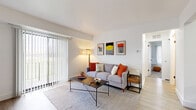 This single-floor residence offers a well-organized layout with a clean, contemporary aesthetic carried throughout each room. The living area serves as the heart of the home, featuring light wood-style flooring, white walls, and a large sliding glass door fitted with vertical blinds that floods the space with natural light and provides direct access to an outdoor balcony. A bold accent wall near the entry, dressed in a retro-inspired orange patterned wallpaper, adds a striking focal point and visual warmth upon arrival.

The galley-style kitchen is efficiently designed with white shaker cabinetry featuring black hardware, white countertops, a stainless steel sink with a matte black faucet, and stainless steel appliances including a refrigerator, range, and over-the-range microwave. Light wood-style flooring continues here, maintaining visual cohesion.

The primary bedroom is a comfortable retreat with plush carpeting, neutral walls, and a generous walk-in closet. A large window brings in ample natural light. The secondary bedroom also features carpet flooring and an open closet with shelving. The bathroom includes a full soaking tub with white tile surround and chrome fixtures, offering a clean, classic finish.