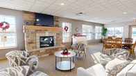 Located at The Estates in Moorhead, Minnesota, this single-floor lobby and community space serves as a central gathering hub for residents and guests. The space is thoughtfully divided into distinct functional zones, all unified by consistent neutral-toned carpeting, a drop tile ceiling with recessed lighting, and warm wood trim accents throughout.

The lounge area is anchored by a striking floor-to-ceiling natural stone fireplace, complete with a wood mantel and a wall-mounted flat-screen television above. Comfortable patterned armchairs and a round coffee table are arranged to encourage conversation and relaxation. Multiple windows along the exterior wall flood the space with natural light and provide views to the outdoors.

Adjacent to the lounge, a dedicated dining area features several round and rectangular wooden tables with matching chairs, positioned near additional windows that continue to bring in ample daylight. Exposed wooden beams add a subtle architectural detail to this zone.

A wide, well-lit corridor connects the community spaces and leads residents to the elevator and residential wings beyond. The entryway features a built-in mailbox station, seating nook, and signage, creating a functional and welcoming point of arrival. Decorative framed artwork and potted plants are placed thoughtfully throughout, contributing to the warm, community-oriented ambiance of the space.