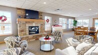 Located at The Estates in Moorhead, Minnesota, this single-floor lobby and community space serves as a central gathering hub for residents and guests. The space is thoughtfully divided into distinct functional zones, all unified by consistent neutral-toned carpeting, a drop tile ceiling with recessed lighting, and warm wood trim accents throughout.

The lounge area is anchored by a striking floor-to-ceiling natural stone fireplace, complete with a wood mantel and a wall-mounted flat-screen television above. Comfortable patterned armchairs and a round coffee table are arranged to encourage conversation and relaxation. Multiple windows along the exterior wall flood the space with natural light and provide views to the outdoors.

Adjacent to the lounge, a dedicated dining area features several round and rectangular wooden tables with matching chairs, positioned near additional windows that continue to bring in ample daylight. Exposed wooden beams add a subtle architectural detail to this zone.

A wide, well-lit corridor connects the community spaces and leads residents to the elevator and residential wings beyond. The entryway features a built-in mailbox station, seating nook, and signage, creating a functional and welcoming point of arrival. Decorative framed artwork and potted plants are placed thoughtfully throughout, contributing to the warm, community-oriented ambiance of the space.