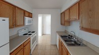 This single-floor unit offers a straightforward, functional layout designed for comfortable everyday living. The galley-style kitchen is well-equipped with warm oak cabinetry lining both walls, providing ample upper and lower storage. A double-basin stainless steel sink, a white electric range with overhead microwave, and light tile flooring complete the space, with a hallway leading directly to the unit's entry door.

The living area is generously proportioned, featuring warm beige carpeting, crisp white walls, and a textured ceiling. A large window fitted with vertical blinds allows natural light to fill the room, creating a bright and open atmosphere.

The bedrooms continue the consistent design language of the unit, each finished with the same neutral carpet and white walls. One bedroom includes a standard closet with a full-width hanging rod, offering practical storage. Windows in the bedrooms provide natural light and exterior views.

Throughout the unit, clean white baseboards frame each room, and flush-mount ceiling light fixtures provide steady overhead illumination. The overall floor plan, as seen in the dollhouse view, presents a compact yet efficiently organized single-level layout with a clear separation between the kitchen, living, and sleeping areas.