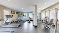 This fitness center offers a thorough and well-organized workout environment designed to accommodate a wide range of exercise needs. The space is laid out across a single level, featuring durable dark rubber flooring throughout that provides both safety and comfort during workouts.

The cardio zone is prominently equipped with multiple treadmills — including Star Trac models — along with elliptical trainers and stationary bikes, offering users a variety of low- and high-impact aerobic options. A wall-mounted flat-screen television is positioned near the treadmill area, enhancing the cardio experience. Large sliding glass doors and windows in this zone flood the area with natural light and provide views of the surrounding outdoor landscape.

The strength training section houses several cable and weight machines, including multi-functional cable systems and weight-stack resistance equipment, well-suited for both isolated and compound exercises. Structural columns divide the space functionally without obstructing movement between zones.

The walls are painted in a calming combination of soft teal and warm beige tones, accented by motivational artwork and sports-themed wall décor. Recessed ceiling lighting ensures consistent illumination throughout. The overall ambiance is clean, functional, and welcoming, making this fitness center a practical and appealing facility for daily exercise.