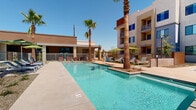This expansive outdoor amenity space is designed to deliver a resort-style experience within a contemporary residential community. The centerpiece is a generously sized rectangular swimming pool with crystal-clear blue water, complemented by a separate spa/hot tub for relaxation. The pool deck is finished in smooth concrete and provides ample space for sunbathing, featuring a row of modern chaise lounge chairs arranged along the poolside.

A standout architectural element is the large pergola-style shade structure, which offers a covered outdoor retreat furnished with additional lounge seating and tables, making it ideal for socializing or simply escaping the sun. Vibrant green market umbrellas are positioned throughout the deck, adding both color and practical shade coverage.

Tall, mature palm trees are strategically placed around the space, reinforcing the warm, desert-climate aesthetic and providing a sense of lush, resort-like character. The surrounding multi-story building features a modern stucco exterior with bold accent colors, large windows, and private balconies that overlook the pool area. The entire amenity space is enclosed by a low metal fence, ensuring a secure and defined recreational zone. Accessibility features, including a pool lift, are also present, reflecting an inclusive design approach.