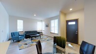 This single-floor space showcases a thoughtfully designed open-concept layout that seamlessly connects the living and dining areas. The living room is anchored by a tufted dark blue sofa and a coordinating accent chair, arranged atop a vibrant geometric-patterned area rug in tones of teal, gold, and grey that adds a contemporary focal point to the space. Light hardwood-style flooring runs throughout, contributing to a warm, cohesive aesthetic.

Multiple windows with white horizontal blinds line the walls, allowing generous natural light to fill the interior and creating an airy, welcoming atmosphere. Clean white walls and smooth ceilings with recessed lighting further enhance the brightness of the space.

The dining area, positioned near the entryway, features a glass-top table paired with sleek dark chairs, adding a modern, sophisticated touch. A decorative grass plant in a dark container provides a natural accent near the entry. The front door, finished in a dark tone with a transom window above, offers an elegant first impression. Overall, the space presents a clean, modern environment well-suited for comfortable everyday living.