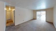 This single-floor, two-bedroom, one-and-a-half-bathroom residence offers a well-organized layout designed for comfortable everyday living. The open living area features neutral-toned carpet, clean white walls, and a sliding glass door fitted with vertical blinds that invites natural light and provides access to the outdoors. A ceiling-mounted light fixture illuminates the entry area, which flows seamlessly into the main living space. The kitchen is outfitted with rich, dark-stained wood cabinetry that extends to the ceiling for generous storage, complemented by light-toned countertops, a double-basin sink, and white appliances including a refrigerator, dishwasher, and range. Both bedrooms are carpeted and feature bright white walls, with windows allowing in natural light and closet access for storage. The full bathroom includes a tiled tub-and-shower combination with a curved curtain rod, set alongside a vanity with a wide mirror, a granite-style countertop, and bright overhead lighting. The half bathroom offers a similarly clean and functional design with a single vanity sink and matching fixtures. Throughout the unit, brushed nickel hardware and neutral finishes create a cohesive, understated aesthetic.
