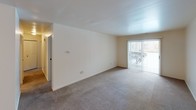 This single-floor, two-bedroom, one-and-a-half-bathroom residence offers a well-organized layout designed for comfortable everyday living. The open living area features neutral-toned carpet, clean white walls, and a sliding glass door fitted with vertical blinds that invites natural light and provides access to the outdoors. A ceiling-mounted light fixture illuminates the entry area, which flows seamlessly into the main living space. The kitchen is outfitted with rich, dark-stained wood cabinetry that extends to the ceiling for generous storage, complemented by light-toned countertops, a double-basin sink, and white appliances including a refrigerator, dishwasher, and range. Both bedrooms are carpeted and feature bright white walls, with windows allowing in natural light and closet access for storage. The full bathroom includes a tiled tub-and-shower combination with a curved curtain rod, set alongside a vanity with a wide mirror, a granite-style countertop, and bright overhead lighting. The half bathroom offers a similarly clean and functional design with a single vanity sink and matching fixtures. Throughout the unit, brushed nickel hardware and neutral finishes create a cohesive, understated aesthetic.