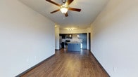This single-floor unit is designed with an open-concept layout that seamlessly connects the kitchen and living areas, creating a spacious and functional environment. The kitchen is well-appointed with dark cabinetry, stainless steel appliances including a refrigerator, microwave, and range, and light granite countertops that provide a striking contrast. A peninsula with a pendant light above serves as a natural dividing point between the kitchen and living space, offering additional counter seating and prep space. A small chandelier above the kitchen adds a refined touch to the area.

The living space is generously sized, featuring rich hardwood-style flooring that extends throughout the unit. A ceiling fan is centrally positioned, offering both lighting and air circulation. Neutral gray walls provide a clean, versatile backdrop suitable for a variety of décor styles. A window near the entryway allows natural light to filter into the space, brightening the interior. The dark front door adds a bold, contrasting design element. The overall ambiance is modern and practical, balancing everyday functionality with contemporary finishes.