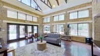 This single-floor gathering space makes an immediate impression with its dramatic vaulted ceilings supported by exposed wood and steel beams, lending a rustic yet modern architectural character. The interior is defined by floor-to-ceiling natural stacked stone walls that carry throughout the space, providing a warm, textured backdrop that ties each area together cohesively.

The main living and lounge area is generously proportioned, featuring rich hardwood flooring, a comfortable gray sofa, and a decorative coffee table set atop a patterned area rug. An array of oversized windows spans multiple levels of the stone walls, flooding the interior with natural light and offering views of the surrounding mature trees and outdoor patio areas. A wall-mounted flat-screen television and open shelving units add functional versatility to the lounge.

A dedicated dining area includes a distinctive herringbone-patterned wooden round table accompanied by woven rattan chairs. French doors open directly to an outdoor patio, seamlessly extending the space for gatherings or events.

The fully equipped kitchen features warm maple cabinetry, granite countertops, stainless steel appliances including a range, built-in microwave, and dishwasher, all framed by stone archways that connect it to the main living area. Stone-tiled flooring in the kitchen complements the overall design aesthetic, creating a cohesive and welcoming environment throughout.