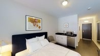 This single-floor studio unit offers a thoughtfully designed living space that combines comfort and functionality in a clean, modern aesthetic. The sleeping area is anchored by a generously sized bed dressed in crisp white linens, accompanied by a cushioned accent chair — creating a cozy and inviting atmosphere. The walls are finished in a neutral white palette, lending the space a bright and airy feel, while framed artwork adds a tasteful decorative touch.

Integrated seamlessly into the same open layout is a compact kitchenette, outfitted with white cabinetry, a dark countertop, a stainless steel sink, a microwave, and a compact refrigerator. This self-contained setup provides essential conveniences for everyday living or extended stays without the need for a separate kitchen space.

The flooring throughout features dark hardwood, providing a grounding contrast to the light walls and adding a sense of warmth to the overall design. Recessed and flush-mount ceiling lighting ensures the space is well-illuminated throughout. The overall ambiance is clean, minimal, and functional — well-suited for individuals seeking a practical yet comfortable living or lodging environment.