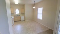 This single-floor space offers a bright, open layout that seamlessly integrates the kitchen and dining area. The kitchen is equipped with light wood cabinetry, a white dishwasher, and a white range, complemented by a distinctive oval window above the sink that allows natural light to fill the space. The countertops and cabinetry maintain a clean, neutral aesthetic that contributes to the room's airy feel. The dining area flows naturally from the kitchen and is distinguished by a classic pendant light fixture suspended from the ceiling, adding a focal point to the space. A standard window fitted with blinds on the adjacent wall further brightens the area. Light-toned tile flooring extends throughout both areas, providing a cohesive and easy-to-maintain surface. The walls are finished in a soft, neutral beige tone, enhancing the overall sense of openness. White baseboards provide a crisp contrast against the flooring. The layout is practical and straightforward, making efficient use of the available space while maintaining a clean, functional design suited for everyday living.