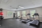 Located at 701 Baywood Cir in Morrow, Georgia, this single-story fitness center offers a spacious and well-organized workout environment designed to accommodate a variety of exercise routines. The facility is laid out across an open, L-shaped floor plan, providing ample room for both cardio and strength training activities without feeling crowded.

The flooring throughout is durable rubber gym matting in a dark speckled finish, well-suited for high-traffic fitness use. Neutral beige walls are accented by a continuous chair rail detail, lending a clean and polished appearance to the space. Textured ceilings are fitted with flush fluorescent light fixtures and ceiling fans, ensuring consistent lighting and air circulation throughout.

Multiple windows fitted with white horizontal blinds line the walls, drawing in generous natural light and offering views of the surrounding greenery. The cardio zone is equipped with multiple treadmills and an elliptical trainer, while a stationary spin bike and rowing machine provide additional low-impact options. The strength training area includes a cable machine, a weight bench, a dumbbell rack with a full set of free weights, and additional resistance equipment. The facility connects to an adjacent room, adding further functional versatility to the overall space.