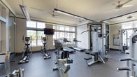 This fitness center is a comprehensively outfitted single-level facility designed to support a full range of workout routines. The space is bright and airy, with large multi-pane windows lining multiple walls and allowing abundant natural light to flood the interior. Views through the windows reveal a pool area and lush palm trees, creating an inviting outdoor connection during workouts.

The cardio zone includes multiple treadmills, elliptical trainers, and stationary bikes, all positioned to take advantage of the outdoor views. Wall-mounted flat-screen televisions are strategically placed throughout the space for entertainment during exercise sessions. Overhead, multiple ceiling fans provide airflow, complemented by linear fluorescent lighting fixtures that ensure consistent illumination throughout the facility.

The strength training area is well-stocked with a variety of cable and selectorized weight machines, a full dumbbell rack, exercise balls, and dedicated floor space with rubber matting for functional training. Floor-to-ceiling mirrors along one wall visually expand the space and allow for form monitoring. The durable rubber flooring throughout provides safety and sound absorption. The overall ambiance is clean, open, and functional, with a layout that comfortably accommodates simultaneous users across all training zones.