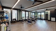 This single-level fitness studio is thoughtfully designed to support a variety of workout routines in a polished, modern setting. The open floor plan is anchored by hardwood-style flooring that runs throughout, providing a clean and durable surface ideal for both cardio and strength training activities. A row of spin bikes is arranged along the center of the room, offering ample space for cycling classes or individual sessions. Floor-to-ceiling mirrors line one wall, visually expanding the space and allowing users to monitor their form during exercise. A large flat-screen television is mounted above the mirror, adding an entertainment and instruction element to the room. Natural light flows in through multiple windows fitted with blinds, creating a bright and welcoming atmosphere. Ceiling fans are installed overhead, contributing to air circulation and comfort during intense workouts. The free weight area features well-stocked dumbbell racks along with medicine balls and resistance equipment, accommodating a wide range of strength training needs. Dark trim accents frame the walls and ceiling, providing a sharp contrast against the sage-green walls and adding a refined aesthetic to the space. Recessed linear lighting supplements the natural light, ensuring the studio remains well-lit at all times. The result is a comprehensive, attractively designed fitness facility suited for diverse training goals.
