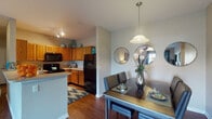 This thoughtfully designed single-floor residence offers a cohesive layout that balances functionality and comfort throughout. The kitchen is a standout feature, equipped with warm honey-toned cabinetry, generous counter space, a double sink, and black appliances including a range, built-in microwave, and refrigerator. Track lighting illuminates the space, and rich hardwood-style flooring ties the kitchen seamlessly into the adjacent dining area. The dining space is defined by a stylish pendant light and a trio of decorative circular mirrors, creating an elegant yet intimate setting for meals. The living room offers a warm, inviting atmosphere with soft ambient lighting, hardwood-style floors, and neutral wall tones that lend a calming backdrop. The bedroom is carpeted for comfort and features a ceiling fan, large windows that invite natural light, and access to a spacious walk-in closet outfitted with wire shelving systems for ample storage. The bathroom is sleek and functional, featuring a glass-enclosed shower, vanity with countertop sink, and hardwood-style flooring. A dedicated in-unit laundry room accommodates a full-size washer and dryer alongside additional shelving, completing this well-rounded and practical living space.