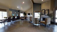 This sophisticated single-floor communal space exudes a modern yet inviting ambiance, designed to serve as a comfortable gathering and resource hub. The interior is anchored by dramatic vaulted ceilings that lend an open, airy feel throughout, with recessed lighting and pendant fixtures providing warm, layered illumination. Large windows allow natural light to filter in, brightening the neutral and deep gray color palette that defines the space's contemporary aesthetic.

At the heart of the room, a sleek fireplace with a refined surround serves as a striking focal point, framed by decorative wall art and flanked by stylish accent chairs upholstered in bold, geometric patterned fabric. A glass-top accent table completes the seating vignette, creating an intimate conversation area. The flooring features large-format stone-look tile, adding texture and visual continuity across the open floor plan.

Toward one end of the space, a dedicated business or computer workstation area is equipped with multiple desktop monitors set along a dark wood counter, ideal for productivity. Nearby, a lounge area with softer furnishings and a wall-mounted flat-screen television provides a relaxed retreat. Potted greenery throughout adds a natural, refreshing element to the polished interior design.