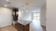 This well-appointed one-bedroom, one-bathroom unit in Greensboro, NC offers a thoughtfully designed open-concept layout that seamlessly connects the kitchen and living areas. The kitchen serves as a central focal point, showcasing rich dark cabinetry, sleek granite countertops, a peninsula island with a double undermount sink, stainless steel appliances including a refrigerator and wall oven, a subway tile backsplash, and a wall-mounted range hood. Pendant lighting above the island adds a modern, refined touch. Wood-style flooring flows throughout the main living area, which is bathed in natural light from a large sliding glass door that opens to a private balcony overlooking the surrounding cityscape. The spacious bedroom features plush carpeting, a ceiling fan, and a large window offering additional urban views, along with direct access to a dedicated closet. The bathroom is finished with light tile flooring, a dark wood vanity, a framed mirror, and brushed nickel hardware, creating a clean, contemporary aesthetic. The overall design strikes a balance between modern finishes and functional living, making this an ideal urban residence in the heart of Greensboro.