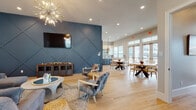 This thoughtfully designed single-floor amenity space blends coastal-inspired aesthetics with modern functionality throughout several distinct zones. Light wood-look tile flooring runs continuously across the entire space, creating a cohesive and airy foundation.

The main lounge area serves as a central gathering point, anchored by a bold slate-blue geometric accent wall adorned with a wall-mounted flat-screen TV. A striking sputnik chandelier overhead adds a sculptural focal point, while a sectional sofa, accent chairs, and woven ottomans create a relaxed seating arrangement. A shuffleboard table nearby provides additional recreational appeal.

Adjacent to the lounge, an open dining area features round wooden tables and chairs positioned near floor-to-ceiling French doors with transom windows above, offering direct views and access to an outdoor pool and patio area. Natural light floods this zone generously.

The kitchen is well-appointed with white upper cabinetry, open shelving, a mosaic tile backsplash, dark lower cabinetry, a center island with a light quartz countertop, stainless steel appliances, and woven pendant lighting overhead.

A separate private meeting or dining room accommodates a large conference-style table with upholstered seating for eight, complemented by pendant lighting, wall art, and a wall-mounted display screen—ideal for formal gatherings or private events.
