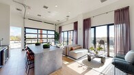 Located at 4312 Yancey Rd in Charlotte, NC, this contemporary single-floor living space is a striking example of modern urban design. The open-concept layout is anchored by a spacious living room that makes excellent use of its footprint through thoughtful design and high ceilings that amplify the sense of space.

Floor-to-ceiling black-framed windows and glass panel garage-style doors flood the interior with abundant natural light, while mauve-toned sheer curtains add a soft, elegant contrast. Light wood-look tile flooring runs throughout, lending warmth and continuity to the space.

A large stone-topped island with bar seating serves as a natural divider, complementing the open living area furnished with a modular sofa, accent chairs, and a sculptural coffee table. One wall features a dramatic bold black-and-white graphic wallcovering, creating a striking focal point anchored by a mounted flat-screen display — an ideal feature for presentations or entertainment.

A private balcony accessed through the glass doors offers open views of the Charlotte skyline and surrounding Yancey neighborhood, bringing the outdoors in. Recessed lighting and in-ceiling speakers complete the refined, contemporary ambiance, making this space well-suited for both relaxation and social gatherings.