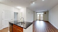 Located at 2300 Woodland Crossing Dr in Herndon, Virginia, this single-floor residence offers a thoughtfully designed open-concept layout that seamlessly connects the kitchen, dining, and living areas. The kitchen serves as a central hub, anchored by a large island with dark granite countertops and a stainless steel sink. Warm-toned wood cabinetry pairs with stainless steel appliances, including a gas range, built-in microwave, and refrigerator, creating a functional and stylish cooking space. Hardwood-style flooring extends throughout the main living areas, enhancing the sense of openness and continuity. The spacious living room is bathed in natural light through glass French doors topped with a transom window, opening to a private balcony with outdoor views. The bedrooms are carpeted for comfort, with generous windows that invite abundant natural light and views of the surrounding neighborhood. Two full bathrooms feature light tile flooring, white vanities with undermount sinks, and large mirrors. One bathroom includes a tub-shower combination with subway tile surround, while the other offers a glass-enclosed shower. The overall aesthetic is clean, neutral, and move-in ready, reflecting a modern yet comfortable residential style.