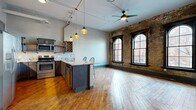 Located in Cleveland's vibrant Ohio City neighborhood, this one-bedroom, two-bathroom loft blends historic character with modern updates. The open-concept living and kitchen area is anchored by original hardwood floors, soaring ceilings with exposed ductwork, and a striking exposed brick wall punctuated by two tall arched windows that flood the space with natural light. The kitchen features dark cabinetry, stainless steel appliances, a built-in wine rack, granite countertops, and a central island with bar seating, all complemented by pendant lighting overhead. A ceiling fan serves the generous living area, enhancing the loft's industrial aesthetic. A long hallway connects the main living space to the private quarters, lined with warm hardwood floors and industrial-style sconce lighting. The bedroom offers a cozy, well-proportioned retreat with hardwood flooring and exposed beam details. Two closets with double doors provide ample storage. Both bathrooms are finished to a high standard, each featuring large-format tile flooring, walk-in glass-enclosed showers with subway and wood-look tile accents, granite vanity tops, dark cabinetry, and rustic reclaimed wood shelving — a thoughtful nod to the building's industrial heritage.