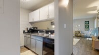 Located at 1117 City Park Ave in Fort Collins, Colorado, this one-bedroom, one-bathroom unit offers a thoughtfully organized layout across a single floor. The open-concept kitchen and living area forms the heart of the home, where wood-style flooring runs throughout and neutral gray walls create a clean, versatile backdrop. The galley-style kitchen is efficiently designed with dual-sided cabinetry in gray, dark granite-style countertops, a stainless steel sink, and full-size black appliances including a range and refrigerator. White subway-style tile lines the kitchen walls, adding a bright, modern touch. The living room flows naturally from the kitchen and features a large window that draws in natural light, wall-mounted floating shelves, and a pass-through counter connecting to a small breakfast bar area. A hallway leads to the private bedroom, which is comfortably sized with plush carpeting, a sliding-door closet for ample storage, and a window allowing natural light. The bathroom includes a full soaking tub and shower combination with clean white tile surround and gray cabinetry with dark countertops, consistent with the home's overall aesthetic. Two hallways and a closet complete the functional layout of this well-appointed Fort Collins residence.