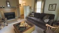 This two-floor home offers a welcoming and well-appointed interior designed for comfortable everyday living. The living room serves as the heart of the space, anchored by a classic fireplace with a wooden mantle that adds warmth and character to the room. Plush, dark upholstered seating surrounds a glass-top coffee table, creating an inviting area for relaxation and conversation. A large window dressed with floor-length curtains allows generous natural light to filter in, brightening the carpeted interior. A decorative folding screen adds a subtle architectural detail, partially framing the transition to an adjacent area. The neutral color palette of warm taupes and soft grays lends a calming, cohesive feel throughout the visible spaces. An area rug with a floral motif adds a touch of pattern and texture underfoot. The layout suggests a thoughtful flow between rooms, with a hallway visible beyond the living area leading to additional spaces across the two floors. Overall, the home presents a traditional and comfortable ambiance, blending functional design with homey, lived-in charm.