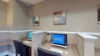 Located at 3280 Mill Run Dr in Hilliard, Ohio, this single-floor business center offers a functional and professional environment designed to meet productivity needs. The space features dedicated computer workstations equipped with desktop computers, keyboards, and mice, each set within individual built-in desk alcoves that provide a sense of privacy and focus. The workstations are finished in a neutral, warm-toned laminate that complements the overall color palette of the room. The walls are adorned with a subtle textured wallcovering in soft beige tones, accented by a white chair rail that adds a refined architectural detail. Decorative canvas artwork featuring abstract designs in muted blues, browns, and creams lends the space a polished, contemporary aesthetic. The layout is compact yet efficient, making practical use of the available floor space to accommodate multiple users simultaneously. The clean lines, neutral tones, and understated décor create a calm, distraction-free atmosphere well suited for business tasks. Overall, the space strikes a balance between functionality and professional design, making it a practical resource for those seeking a dedicated workspace.
