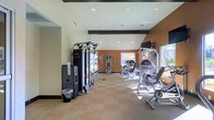 This fitness center offers a comprehensive workout environment designed to accommodate a variety of exercise routines. The space is laid out across a single floor, featuring a spacious open floor plan that allows for easy movement between equipment stations. The interior is characterized by warm terracotta-toned accent walls contrasted against light neutral walls, creating an energizing yet welcoming atmosphere. Recessed lighting is evenly distributed across the ceiling, complemented by natural light streaming in through large windows that provide views of the surrounding greenery outside. The flooring consists of durable, low-pile carpeting in a neutral tone, practical for a high-traffic fitness environment. The room is outfitted with a robust selection of strength-training machines, including cable and pulley systems, multi-station weight machines, and resistance equipment suited for both upper and lower body workouts. Additional cardio and functional training stations are distributed throughout the space. Dark wood beam accents along the upper walls add a subtle architectural detail that lends a polished, finished look to the room. A sanitizing station near the entrance promotes cleanliness and hygiene throughout the facility. Overall, the space balances functionality with a clean, modern aesthetic.