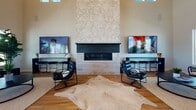 This single-story community clubhouse is designed with a coastal-contemporary aesthetic, combining open, airy interiors with warm, natural finishes throughout. The main lounge area commands attention with dramatically high ceilings and an abundance of natural light streaming in through large windows that frame views of the surrounding neighborhood and a covered outdoor deck. A striking floor-to-ceiling fireplace clad in geometric, textured tile serves as the room's focal centerpiece, flanked by flat-screen TVs, leather seating, and warm wood flooring that runs throughout the space.

The wet bar and kitchenette area features light quartz countertops, wood-toned cabinetry, open floating shelves adorned with potted greenery, and pendant lighting overhead, creating a welcoming gathering spot. Wicker bar stools and an oversized island add to the relaxed, coastal ambiance.

The fitness center is well-appointed with treadmills, ellipticals, free weights, cable machines, and large mirrors, all set atop a distinctive geometric-patterned floor. Natural light pours in through multiple windows, and a ceiling fan keeps the space comfortable.

Wide sliding glass doors and double entry doors open to a spacious covered deck with white railings and rocking chairs, offering views of the pool and community beyond.