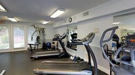 This fitness center is designed to support a comprehensive workout routine within a compact, efficiently arranged space. The room is outfitted with multiple treadmills positioned for easy use, along with a rowing machine and a multi-station cable weight machine, providing options for both cardiovascular and strength training exercises. A large wall mirror spans a significant portion of the room, enhancing the sense of space while allowing users to monitor their form during workouts. The walls are painted in a clean, neutral white, and the textured ceiling is fitted with recessed fluorescent lighting that ensures the room is well-lit throughout. Ventilation is provided through ceiling vents, maintaining a comfortable environment during exercise. A wall clock keeps users on track with their sessions. The flooring is dark and durable, appropriate for a high-use fitness setting. Additional accessories such as exercise balls and free weights are visible, rounding out the available equipment. Overall, the space is practical, organized, and well-maintained, making it a functional facility suited for a variety of fitness needs.